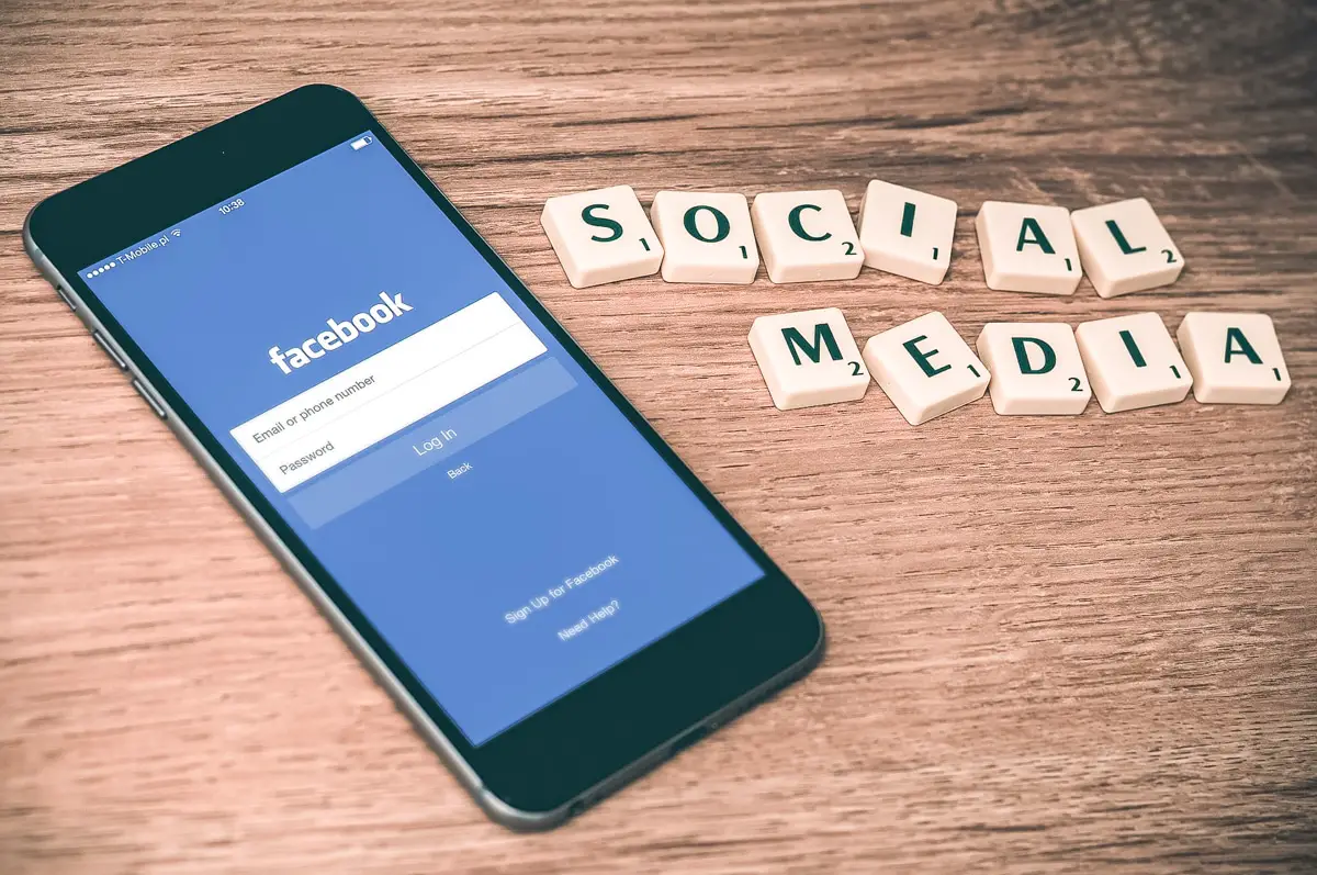 Featured photo for 7 day social media detox showing phone with facebook app opened laying on the wooden table. There's social media sign made from Scrabble blocks beside it