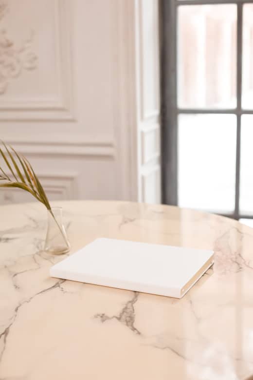 White, plain notebook on the beige table with beige walls and huge floor-to-ceiling window with black frame in the background