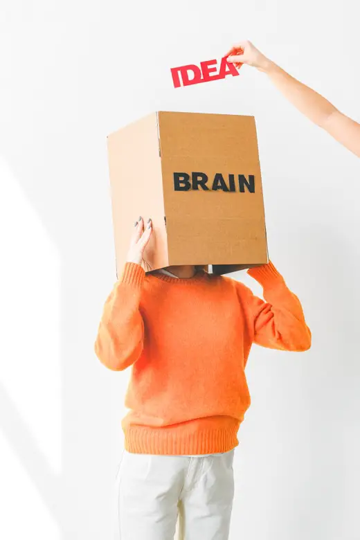 Woman dressed in orange jumper and white jeans keeps her head in the cardboard box with Brain sign on it. Above it you can see someone's hand with Idea sign in it