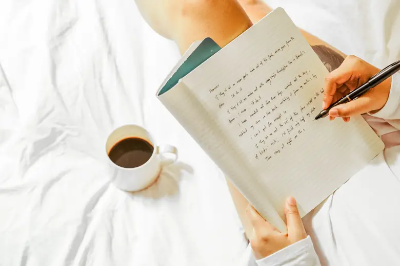 Aerial view of a woman sitting on her bed and writing in her journal, you can see a mug of black coffee next to her - featured image for journaling ideas for beginners