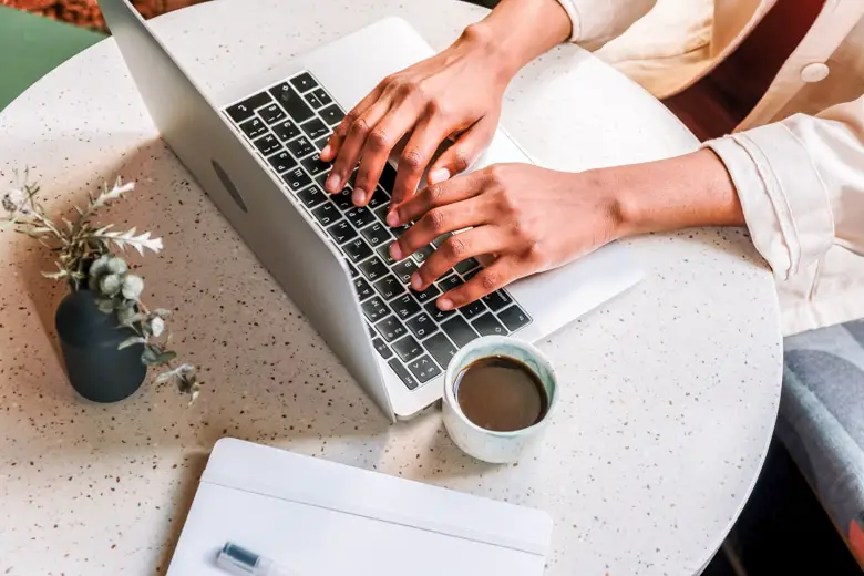 Aerial view of the woman sitting by the white, round table, typing on the laptop with a flower, mug of coffee and a notebook on the side - featured image for blogging advantages and disadvantages