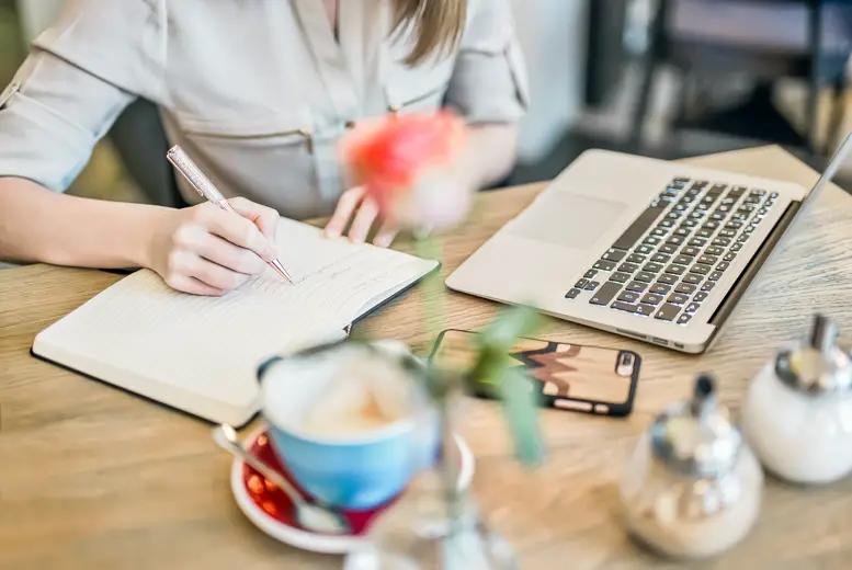 Photo of a woman sitting by a wooden table in a cafe writing in her notebook. There's a laptop, phone and cup of coffee next to her - journaling ideas for beginners