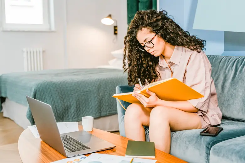 Woman sitting on a coach holding a journal. You can see her laptop sitting on a coffee table in front of her - journaling ideas for beginners