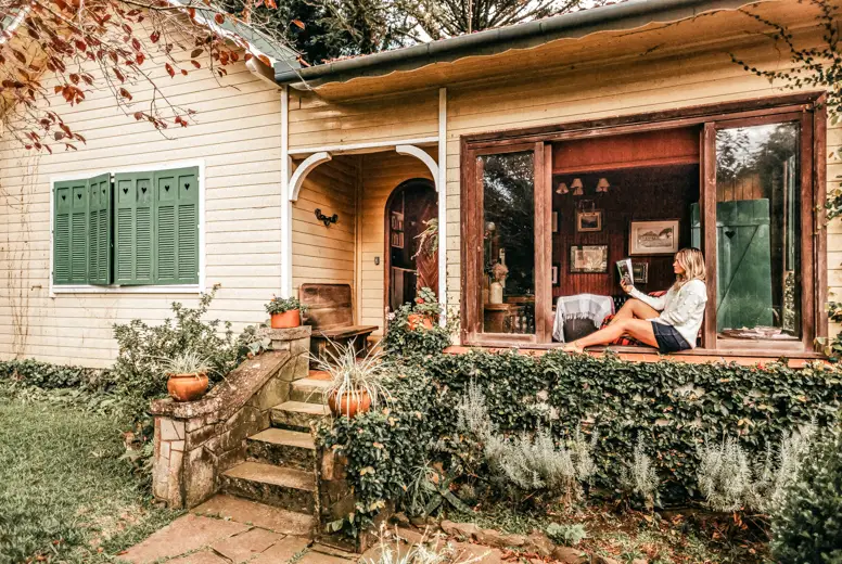 Women sitting on a windowsill and reading a book, she's in an old fashioned, wooden house with a lot of greenery - self care weekend ideas