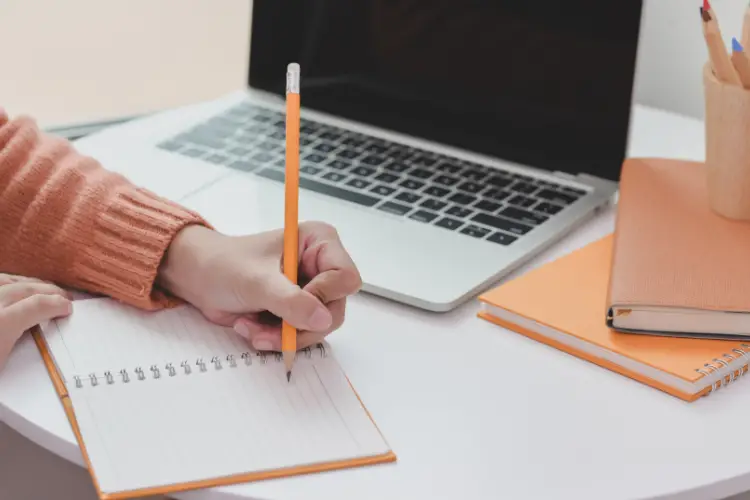 Women writing in a notebook with a pencil sitting by a white table with laptop and notebooks on it - how to reset your life