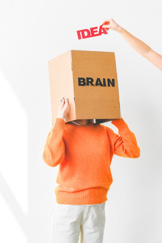 Woman dressed in orange jumper and white jeans keeps her head in the cardboard box with Brain sign on it. Above it you can see someone's hand with Idea sign in it