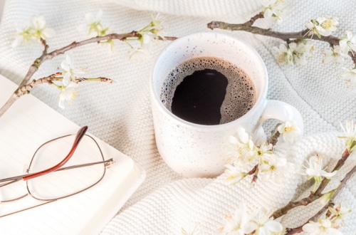 Cup of coffee, glasses, notebook and small branches with flower buds laying on the with blanket - featured image for 9 steps to get your life back on track with mid year reset