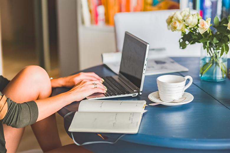 Woman sitting by a round, blue table with a laptop, notebook, and cup of coffee in front of her - featured image for my first blogiversary, 7 things I wish I knew before starting a blog